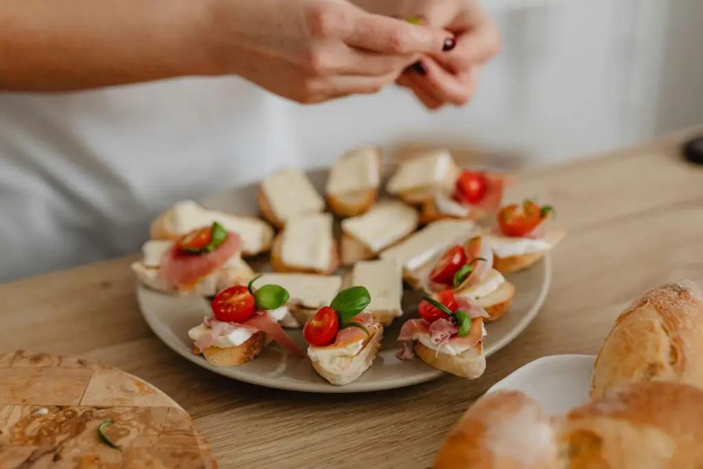 Hands preparing appetizers with cheese, cured meats, and cherry tomatoes on a wooden table.